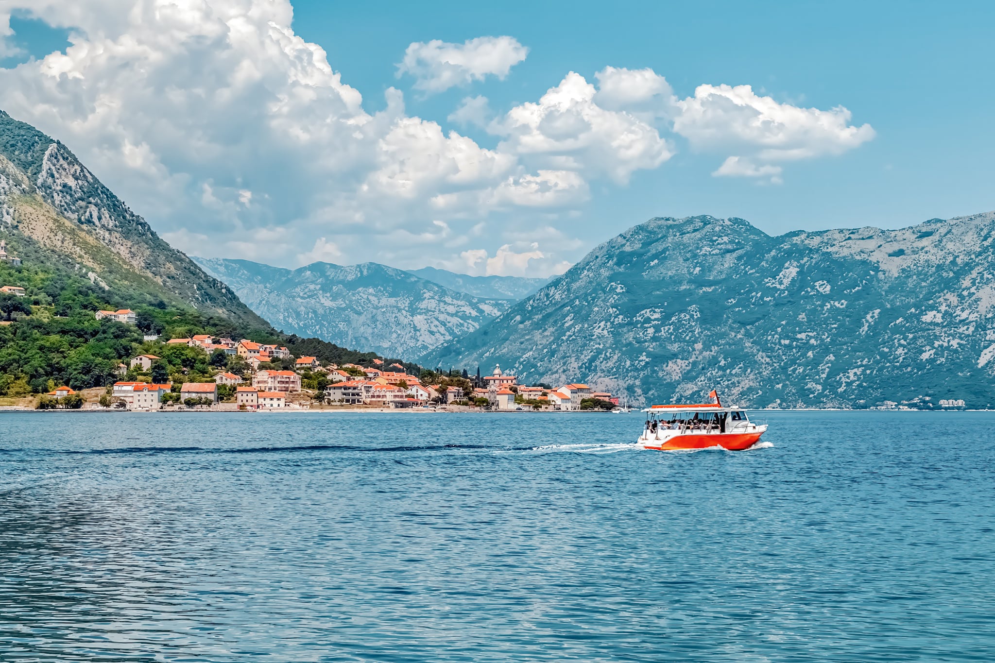 A tour boat sails along the calm waters of Boka Kotorska Bay, passing the picturesque coastal village of Prčanj, Montenegro