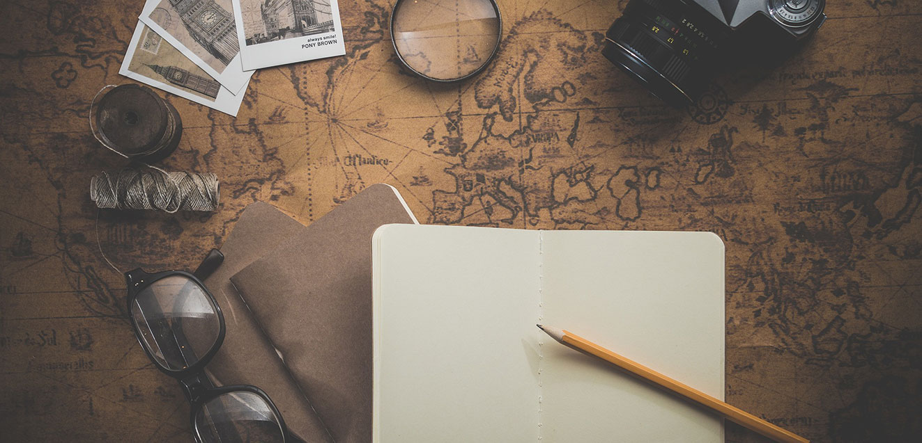 A collection of travel-themed items laid out on an old sepia map. Open notebook and pencil are predominantly featured.