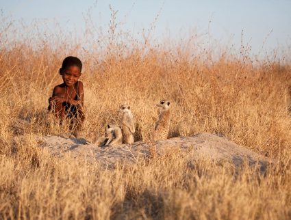 Botswana, a bushman child watches a family of meerkats by the entrance to their burrow.